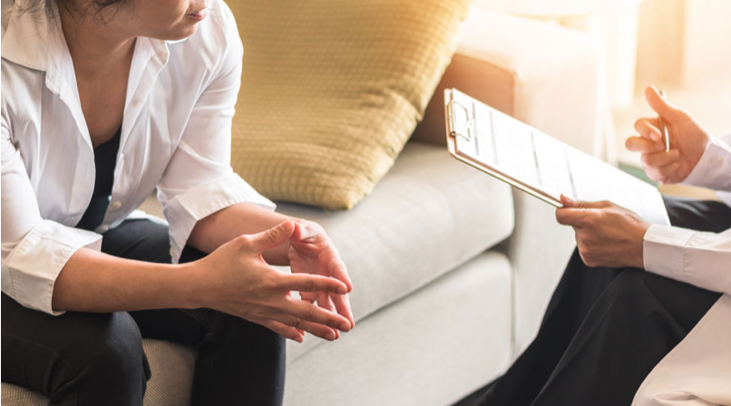 One person on left sits on couch, while therapist sits to right holding notes.